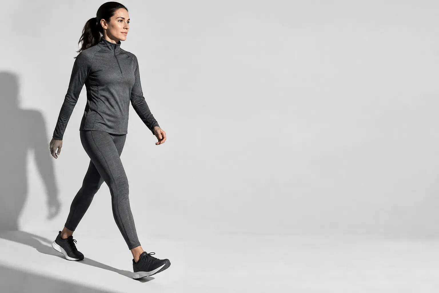 Mujer con conjunto deportivo oscuro caminando con naturalidad sobre un fondo gris claro.