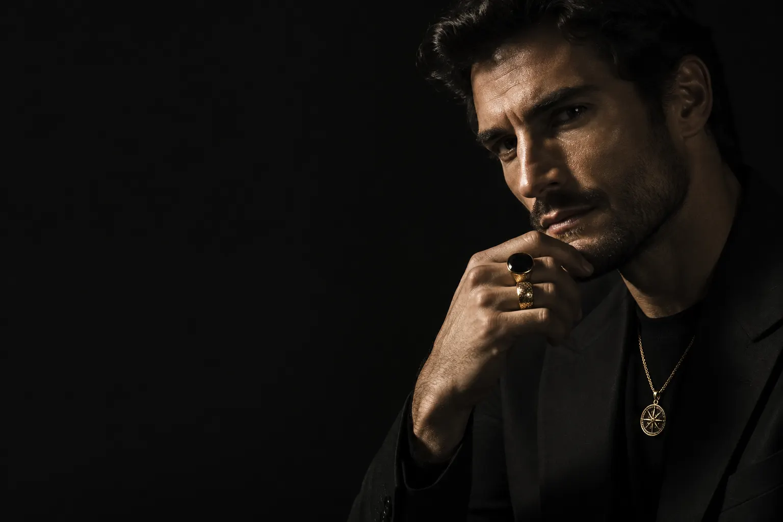 Hombre vestido de negro con anillos y collar, iluminado contra un fondo oscuro.