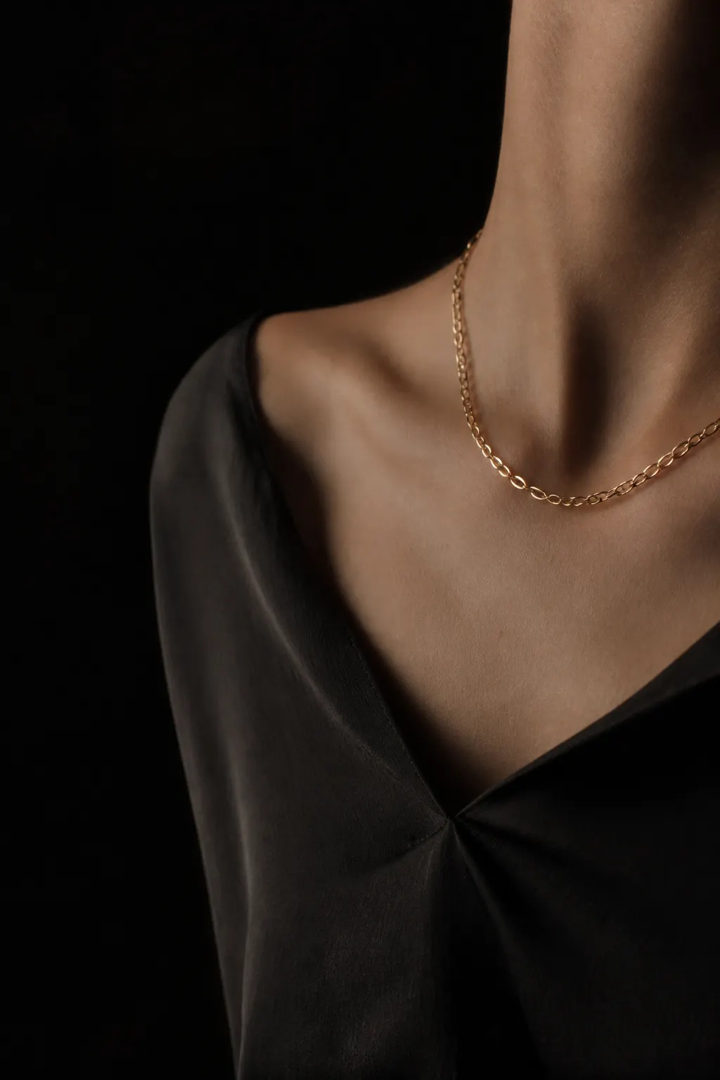Cuello y clavícula sin rostro visible llevando una cadena dorada delicada sobre prenda carbón.