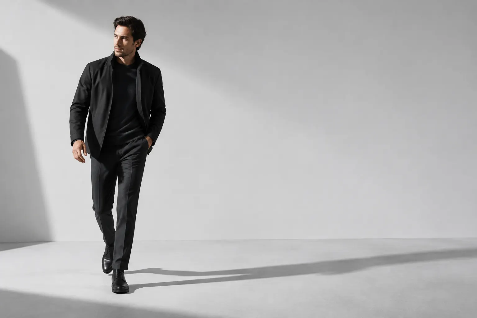 Hombre vestido con chaqueta y pantalón oscuros caminando ante un fondo gris claro.