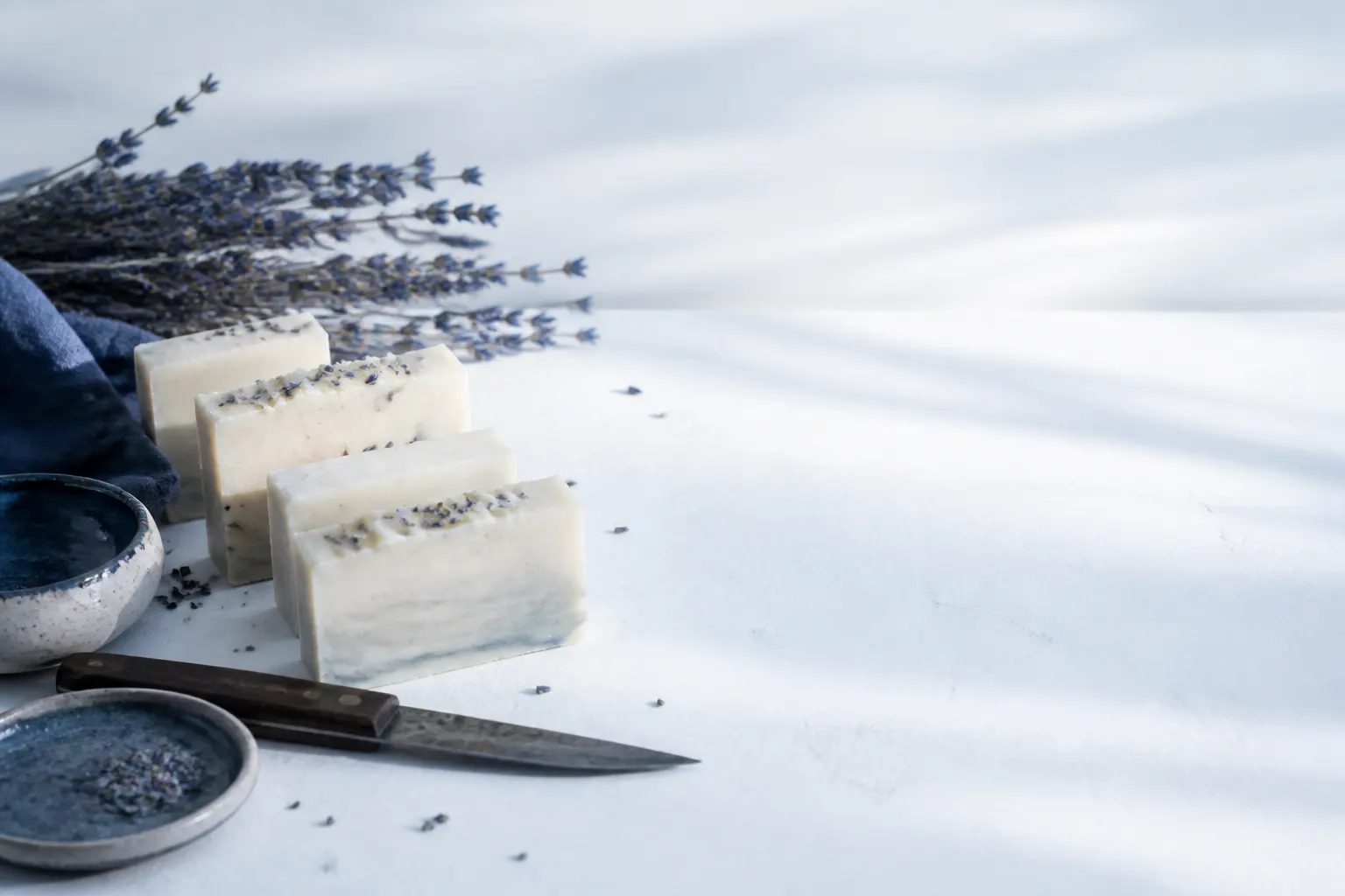 Barras de sabonete artesanal cortadas à mão, lavanda e utensílios de atelier numa bancada clara.