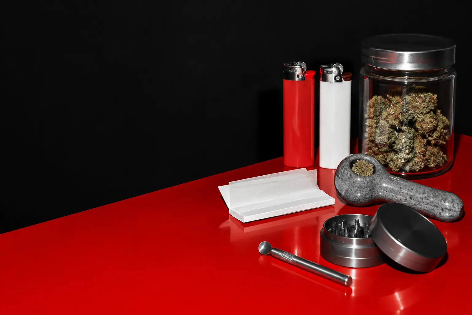 Bodegón de encendedores, papelillos, una pipa y accesorios sobre una superficie roja contra fondo negro.