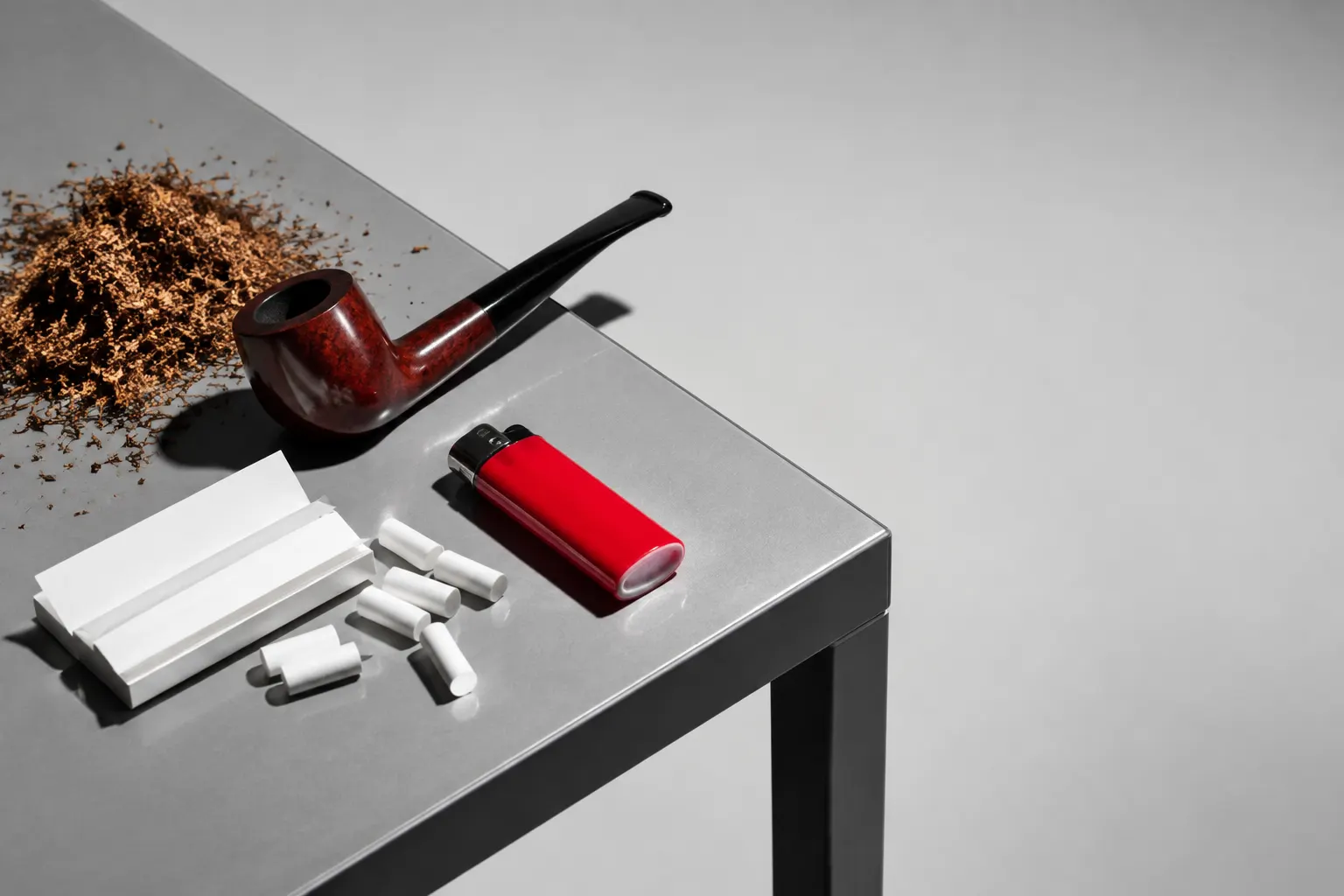 Mesa con tabaco, pipa, papelillos, filtros y encendedor rojo sobre fondo gris claro.