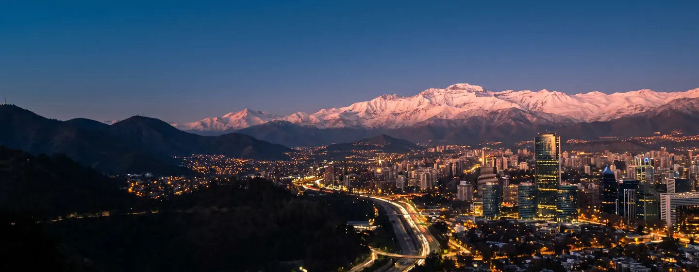 Santiago a la hora azul, con luces de la ciudad, una autopista y la cordillera nevada al fondo.