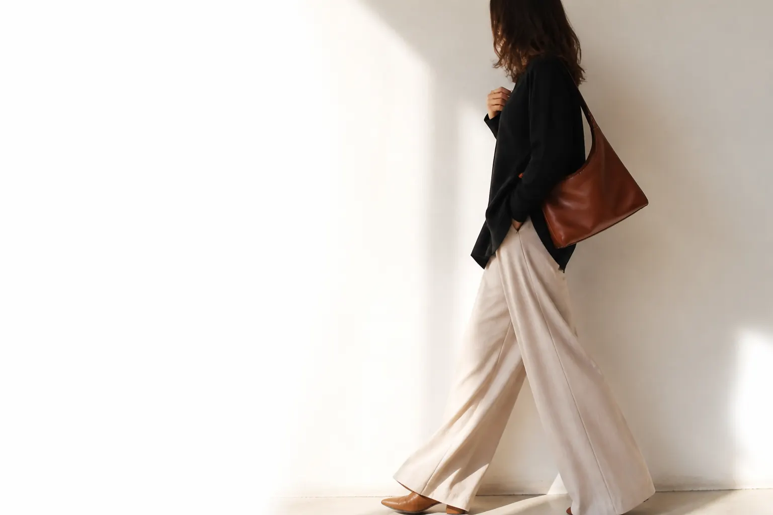 Mulher sem rosto visível, de corpo inteiro, usando um conjunto neutro e segurando uma mala ao ombro contra fundo branco.