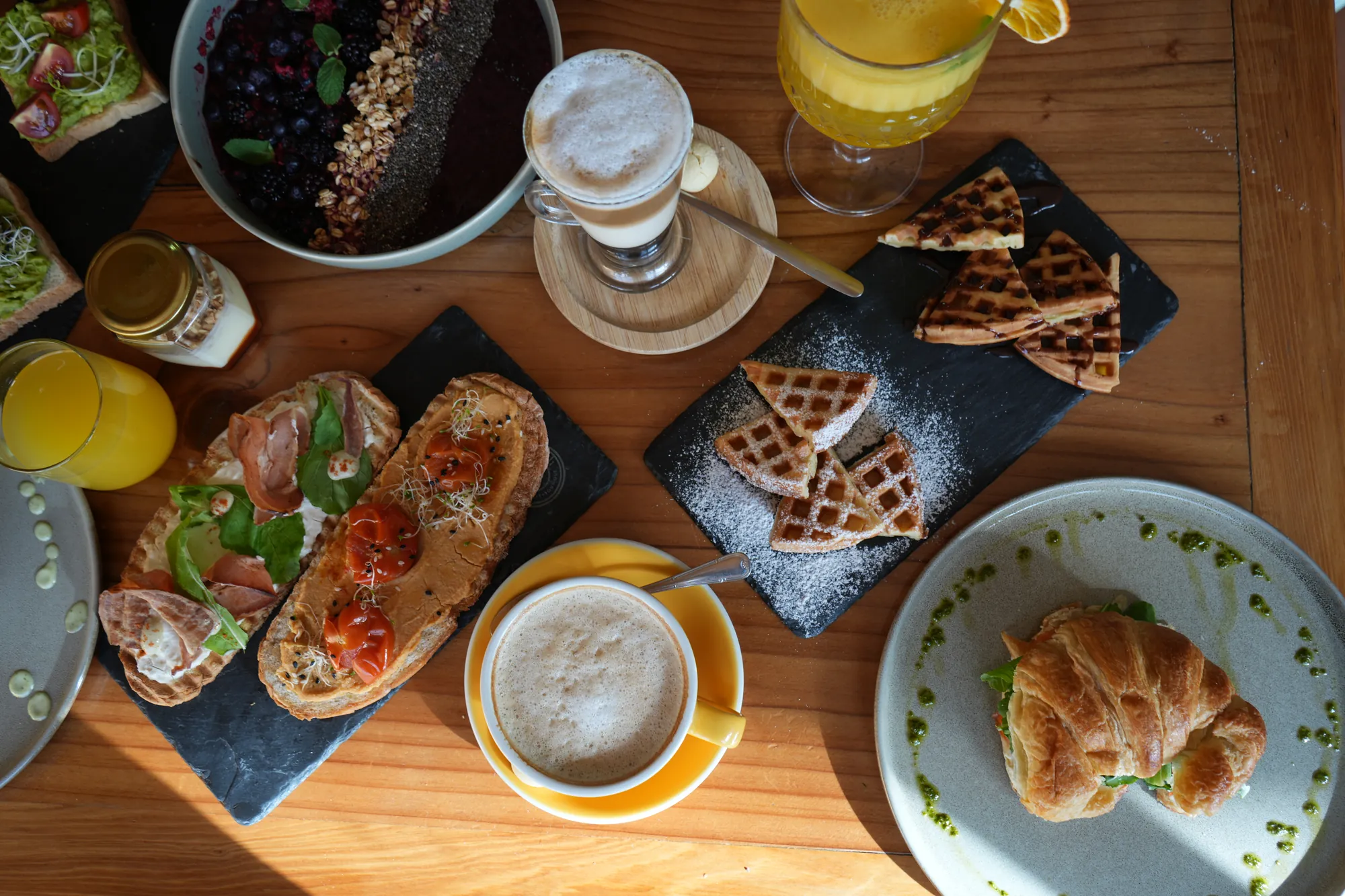 Vista cenital de una mesa de Café Boutique con bowl de açaí, latte, jugo, waffles, tostadas, capuchino y croissant relleno