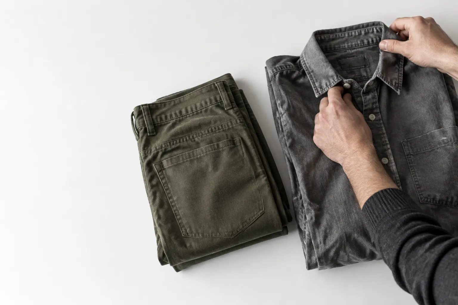 Manos ordenan una camisa y un pantalón masculinos usados sobre una superficie clara.