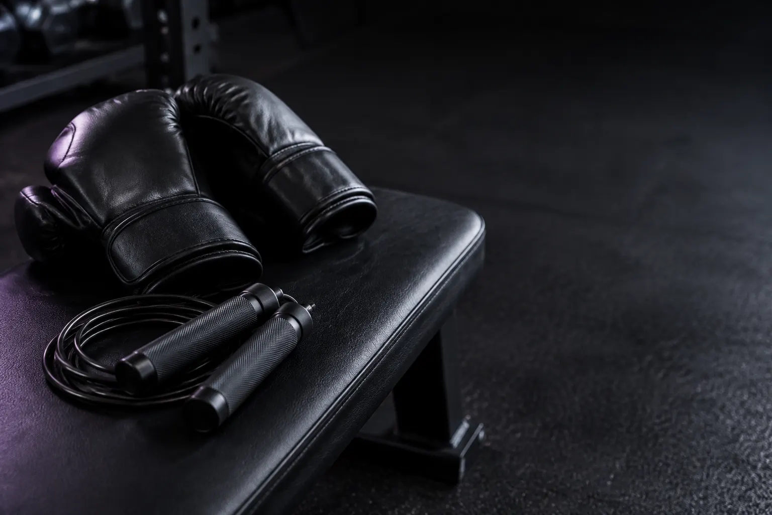 Guantes negros y una cuerda de saltar descansan sobre un banco de gimnasio oscuro.