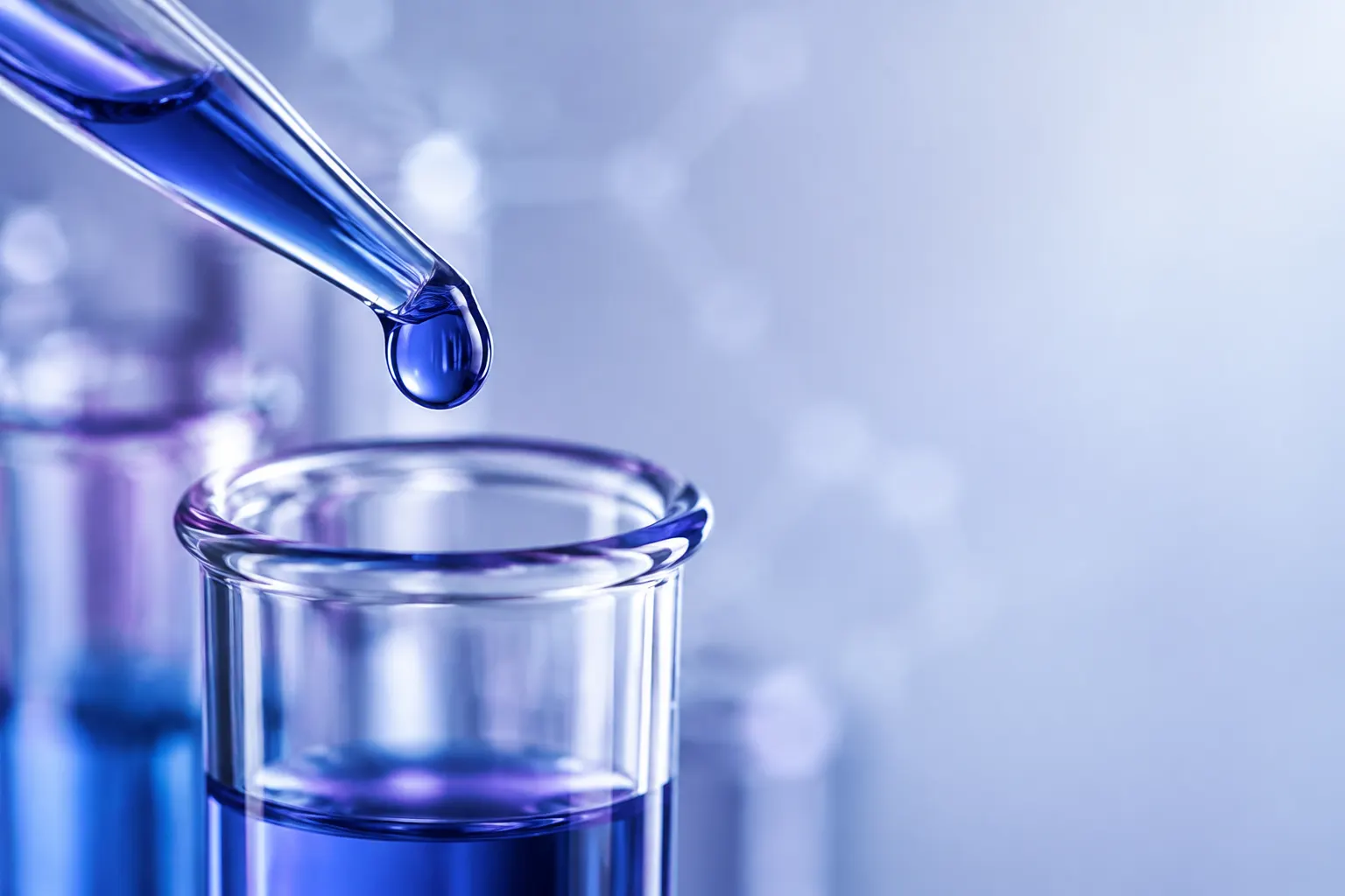 Close-up of a glass pipette and blue droplet beside a clear laboratory vessel.
